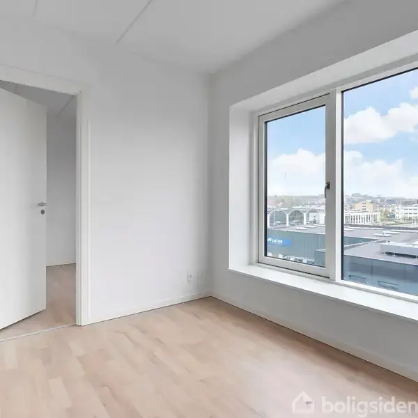 Et tomt værelse med trægulv, åben hvid dør, og stort vindue med udsigt til bygninger og himmel.