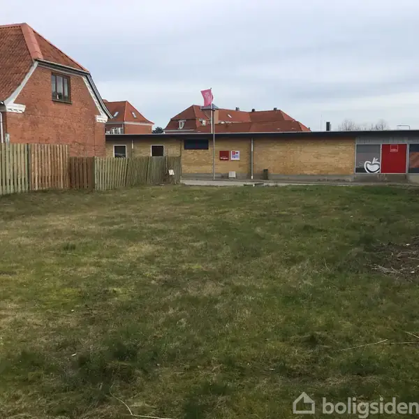 En græsplæne strækker sig mellem et rødt murstenshus til venstre og en lav bygning med cykelsymboler til højre. En træhegn deler plænen fra venstre bygning. Ingen synlig tekst.