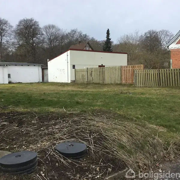 Grundstykke med græs omkranset af hegn og bygninger. Et skilt står foran med teksten "TIL SALG" og kontaktinformation: "68 92 68 11". Baggrunden viser træer og røde bygninger.