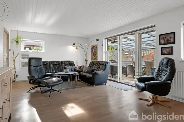 Sort lædersofaarrangement med sofabord står i stue, mens skydedøre fører ud til overdækket terrasse med spisebord. Væggene er hvide, og rummet har trægulv.