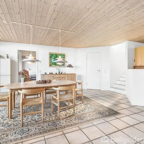 Et spisebord med seks stole står på et tæppe, omgivet af et lyst rum med træloft. Ved siden af er der skænke, kunst på væggen, og et moderne køkken til højre.