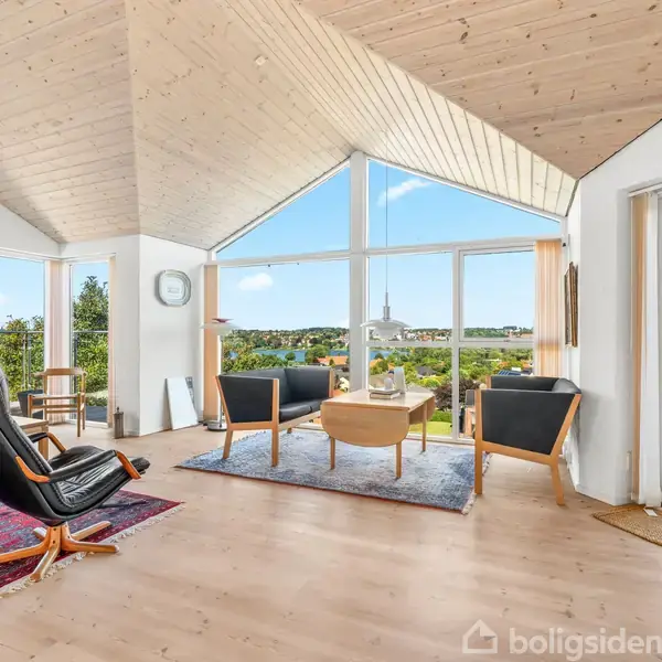 Stue med lænestol og sofagruppe på røde tæpper, vendt mod store vinduer med udsigt over et grønt landskab og blå himmel. Lyst træloft og gulve skaber en varm atmosfære.