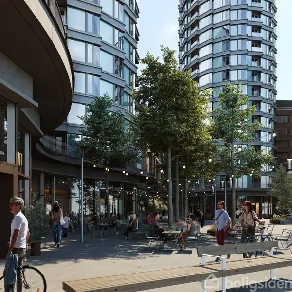 Et moderne byområde med buede boligbygninger omkranser et åbent torv. Folk går eller cykler forbi. På venstre side ses skiltet "post cafe coffee and more". Træer og udendørs caféborde skaber en afslappet atmosfære.