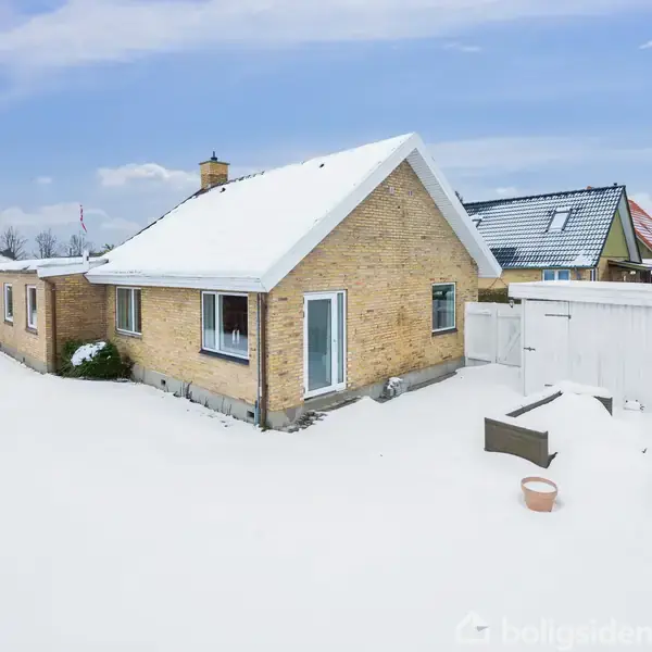 Et gult murstenshus med hvide vinduesrammer står i en snedækket have. Udendørs møbler og et skur ses i baggrunden under en blå himmel med skyer.