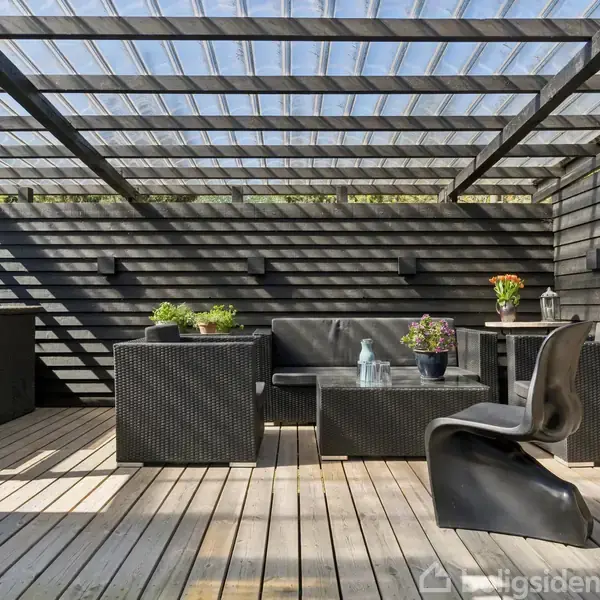 Udendørs terrasse med moderne sorte møbler, herunder sofa, borde og stol. Trægulv og glastag skaber en hyggelig atmosfære. Planter i potter tilføjer grønt.