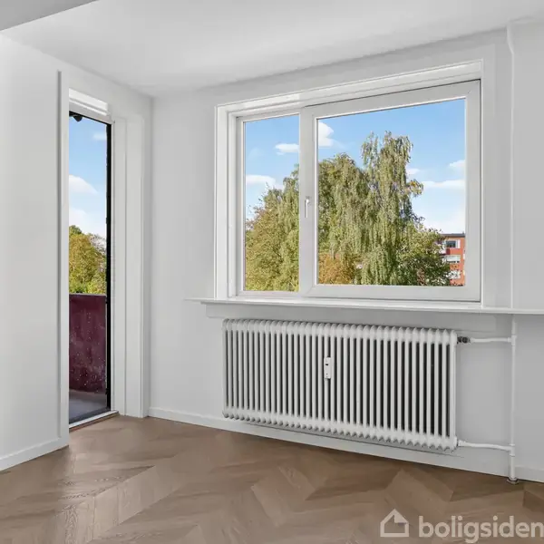 Et lyst værelse med åbent vindue og dør til en lille altan. Radiator under vinduet, udsigt til træer og en bygning i baggrunden.