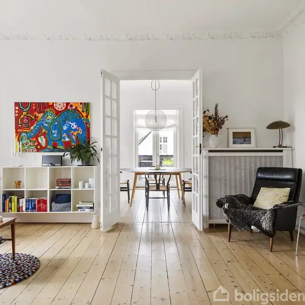 Stue med træplanker, farverig kunst på væggen, sofabord, og lænestol. I baggrunden dobbeltdøre åbner til en spisestue. Hylder indeholder bøger og planter. Skilt "Boligsiden" er til stede.