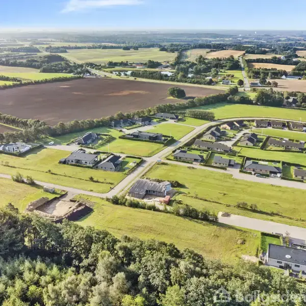 Landskab med nye huse arrangeret i et boligområde. Grønne marker og skov omgiver kvarteret, der er afgrænset af lange, lige veje. Boligerne ligger spredt med store åbne græsplæner.