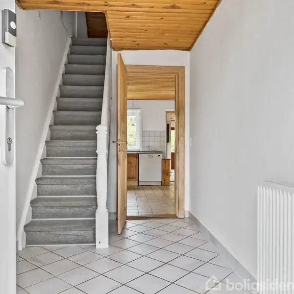 En grå trappe stiger opad, flankeret af hvide vægge. Foran trappen ses en åben dør, der fører til et lyst køkken med træelementer. Flisegulvet dækker entreen.