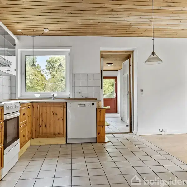 Køkken med træskabe og hvidt flisegulv. Komfuret står ved vinduet, der giver udsigt til træer. To hængelamper hænger fra træloftet, og en dør fører til et andet rum.