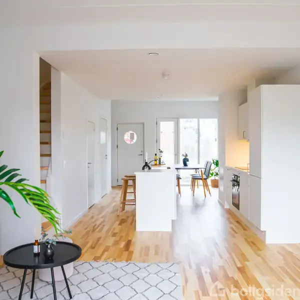 Stue med moderne, lyst interiør. Rundt sofabord foran sofa, åbent køkken med barstole, trægulve, grøn plante og dekorative genstande i naturligt oplyst rum.