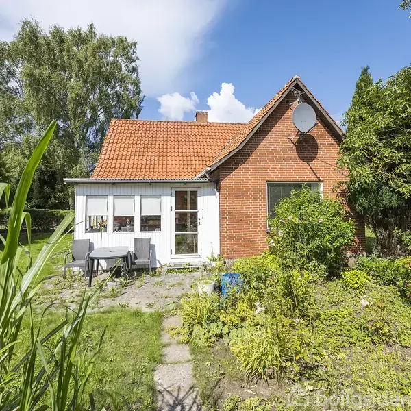 Et rødt murstenshus med rødt tegltag står i en have med grønne planter og blomster. Foran huset er en terrasse med et bord og stole. Solrig dag.