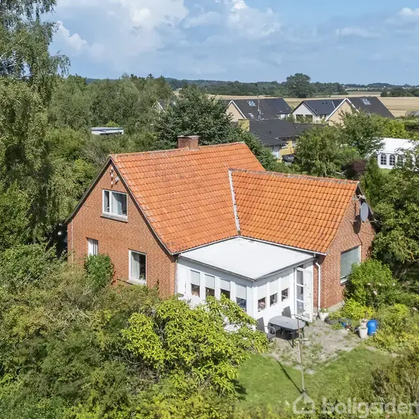Et rødt murstenshus med orange tegltag ligger omgivet af frodig vegetation og træer. Omkringliggende kontekst inkluderer en byvej med et 40 km/t skilt og andre huse i baggrunden.