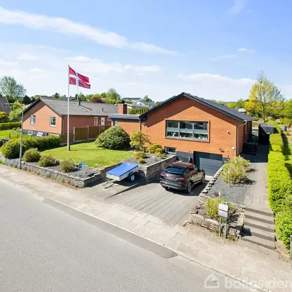 En murstensvilla med fladt tag står i et parcelhuskvarter. Foran huset holder to biler parkeret i indkørslen, og et dansk flag vejrer til venstre under en klar blå himmel.