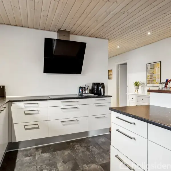 Køkken med hvide skabe og sorte bordplader, vægmonteret sort emhætte. Rummet har trægulv, lyst træloft og hvidmalede vægge. Stegspand og kaffemaskine står på bordet.