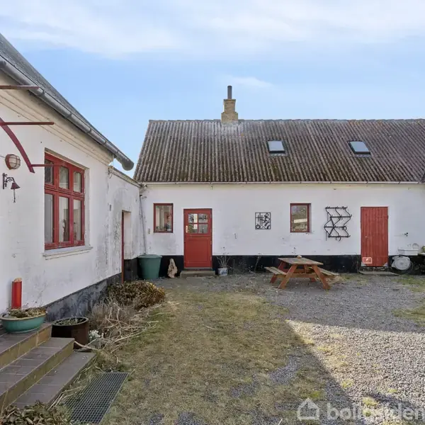 Hvid gårdbygning med røde døre og vinduer. Bord-bænkesæt står i gårdhaven. Omkringliggende græsplæne og grusbelægning under klar blå himmel.