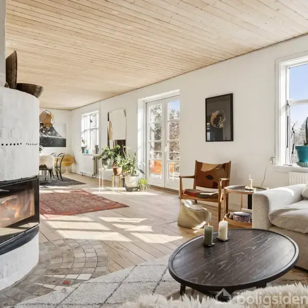 Pejs i hvid stue oplyser rummet, omgivet af sofa og bord med stearinlys. Lyse trægulve og planter skaber en varm atmosfære med store vinduer, der slipper naturligt lys ind.