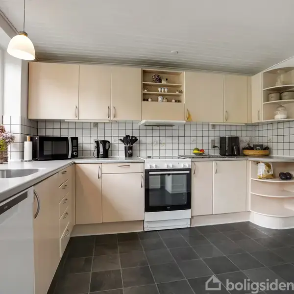 Et lyst køkken med hvide skabe og sorte fliser; komfur og ovn centralt. Mikroovn og elkoger til venstre, glas og skåle til højre på åbne hylder.