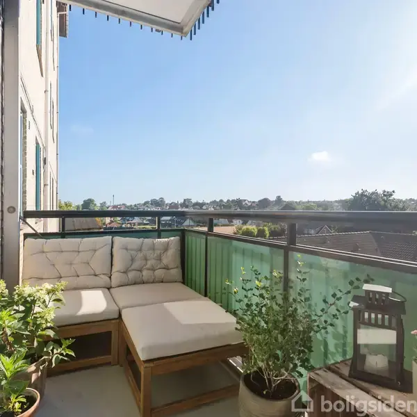 Balkon med beige hjørnesofa placeret mod en mur, omkranset af grønne planter i potter. Udsigt mod en blå himmel og byens hustage.