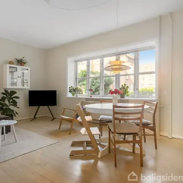 En hyggelig stue med et spisebord og stole nær et stort vindue, der lader sollys ind. En sofa og et TV står til venstre, omgivet af planter.