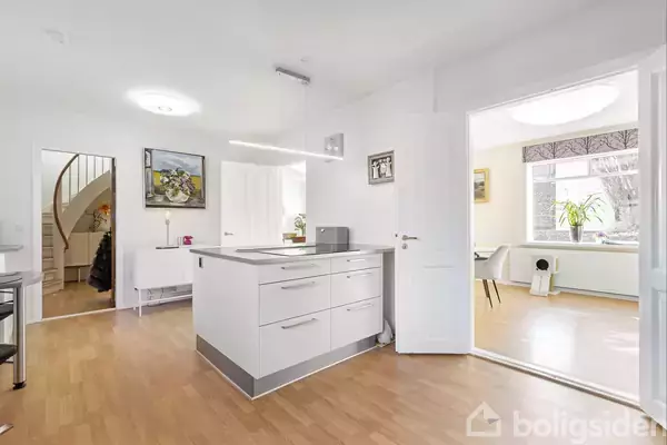 Et lyst køkken med en moderne køkkenø midt i rummet. Bagved er en åben dør, der fører til en stue med et stort vindue. En trappe er synlig i baggrunden.