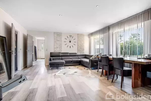 En stue med en stor sort hjørnesofa står mod en væg udsmykket med et ur. Et spisebord med stole er placeret ved et stort vindue med gardiner, der slipper lys ind.