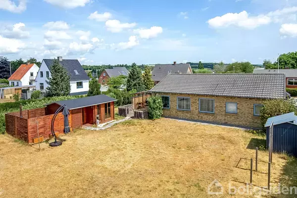 En græsplæne omkranser en gulstensbygning med gråt tag, ved siden af en rød træbygning. Flere huse er synlige i baggrunden under en blå himmel med skyer.