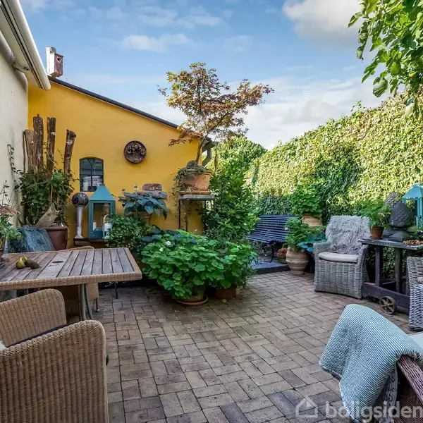 Terrasse med fliser omgivet af grønne planter og hegn. Siddepladser i flet og træbord. Gul murstensvæg bagerst med dekorative elementer og en lille, central plante.