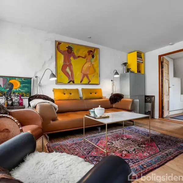 Stue med orange sofa og sofabord på orientalsk tæppe. Malerier og lamper pryder væggene. Døråbning fører til køkken. Lysindfald gennem vinduet giver en hyggelig atmosfære.