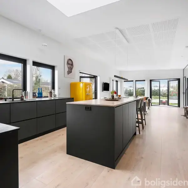 Moderne køkken med sort køkkenø og skabe, gul køleskab, barstole og stort vinduesparti. Rummet er lyst og åbent med udsigt til en have.