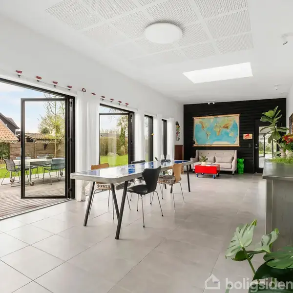 Et lyst værelse med åbne glasdøre fører ud til en have. Indendørs er der et bord med stole, en globus og et vægkort. Plakaten ”Boligsiden” er synlig.