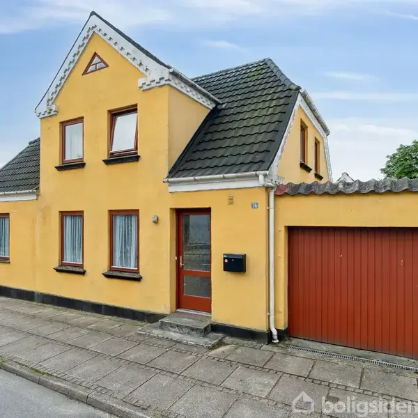 Gul to-etagers hus med røde vinduesrammer og dør, placeret ved en fortovskant. Garagedør til højre. Overdækket med mørkt skråt tag. Blå himmel og træer i baggrunden.