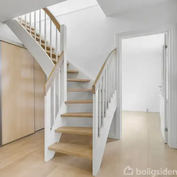 Trætrappe med hvidt gelænder stiger i et lyst rum. Træbeklædte vægge og døre omkranser trappen i et moderne interiør.