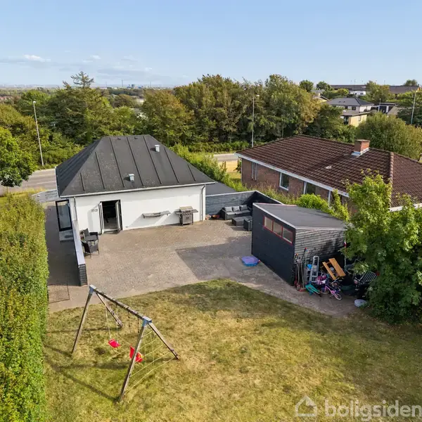 Et hvidt hus med sort tag står stille i en have med græsplæne og gyngestativ. En rummelig terrasse ses ved siden af et mindre skur nær et rødt tegltagshus.