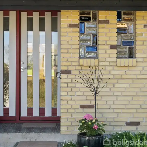 Dør med lodrette glasruder står lukket ved siden af en gul murstensvæg med mosaikvinduer. En potteplante med lyserøde blomster står foran væggen.