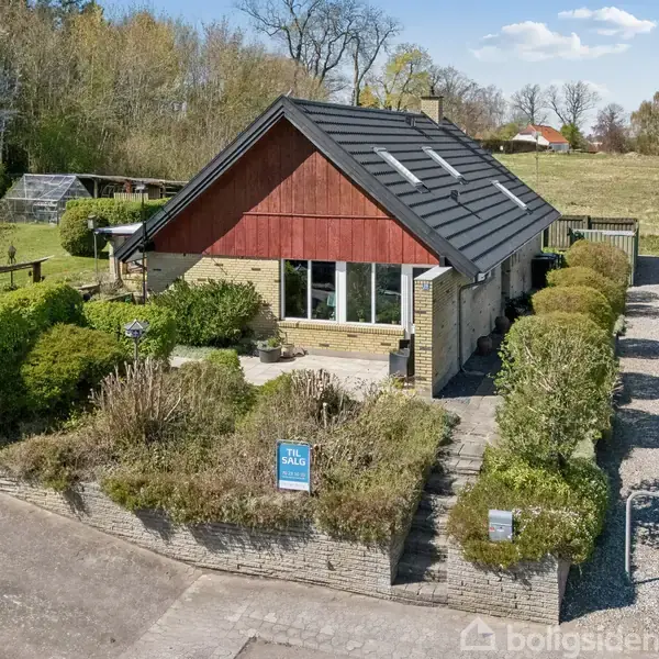Et enfamiliehus med rødt gavlparti og sort tag står i en velplejet have. En grusvej fører forbi, og omgivelserne er grønne med træer i baggrunden. Skilt med "Til salg" foran huset.