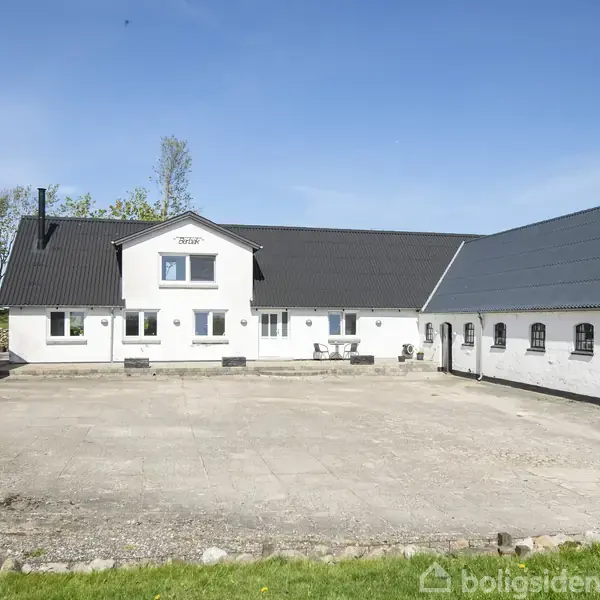 Hvid bygning med sort tag og nummer 7381 på facaden. Bygningen omgiver en gårdsplads med grøn græsplæne forrest. Blå himmel og træer i baggrunden.