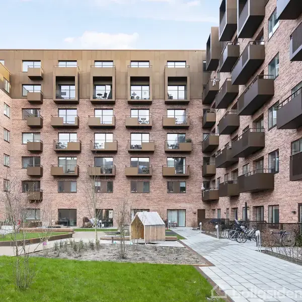 Bygning med røde murstensvægge og mange altaner; omkringliggende gårdhave med græsplæne, stier og cykler står parkeret.