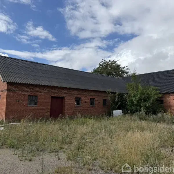 Rød murstensbygning med sorte tagsten står stille. Græsset og ukrudtet vokser vildt omkring bygningen. Himlen er delvis skyet.