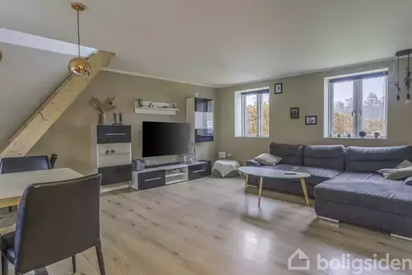 Stue med grå hjørnesofa, sofabord og fladskærms-tv i moderne reol. Spisebord med stole til venstre, trappe opad i hjørnet, lyse træplanker på gulvet, store vinduer.