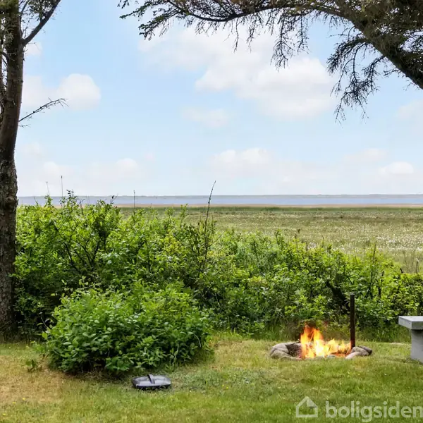 Bål brænder på en græsplæne omkranset af buske og træer, med udsigt over en flad eng mod en blå himmel med spredte skyer. En hængekøje og en bænk står nær bålpladsen.