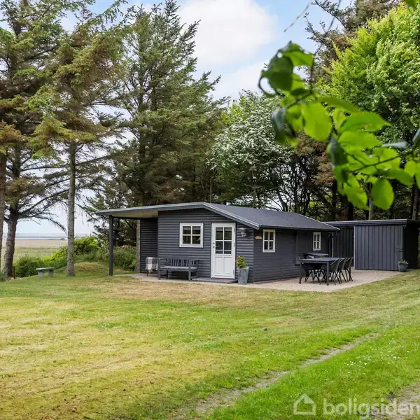Et lille, mørkt træhus står stille omgivet af grønne træer og en stor græsplæne. Der er et sæt udendørsmøbler foran huset, og i baggrunden ses åbent landskab.