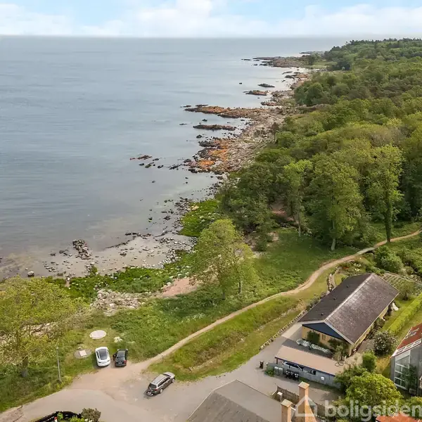 Hus ved kysten på en grøn skovklædt bakke med udsigt til havet og klipper, biler parkeret på en grusvej nær huset. Vandet strækker sig mod horisonten.