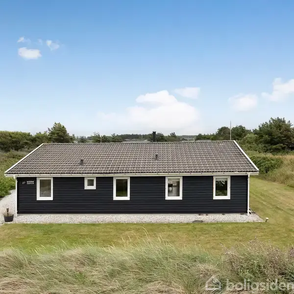 Sort træhus står stille i en grøn have med grussti foran. Omkringliggende buske og træer skaber en rolig, naturskøn atmosfære under en blå himmel med spredte skyer.