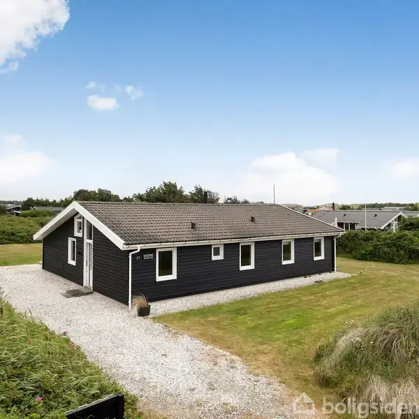 Et sortmalet hus står omgivet af en grusbelagt indkørsel og grønne marker. Det har et gråt sadeltag og flere vinduer mod en lys, blå himmel med spredte skyer.