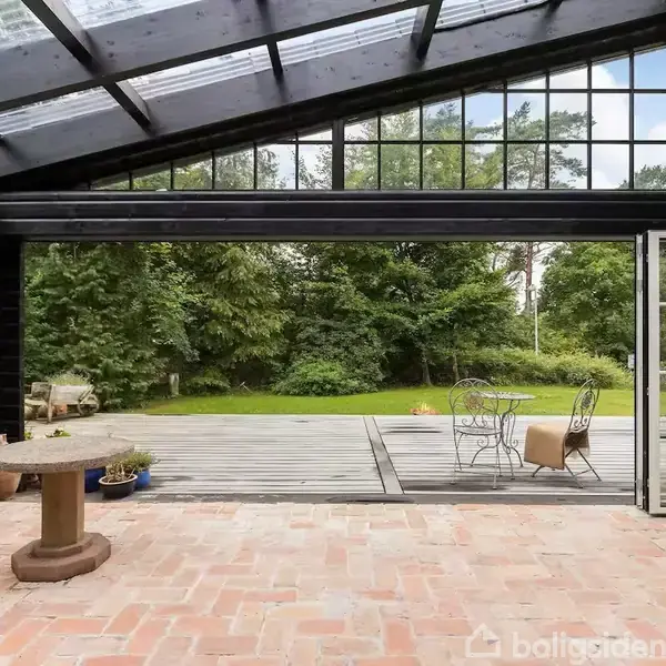 En overdækket terrasse med et glasloft åbner ud mod en have. Terrassedøre står åben, og der ses et bord og stole på trædækket. Skovklædt område i baggrunden.