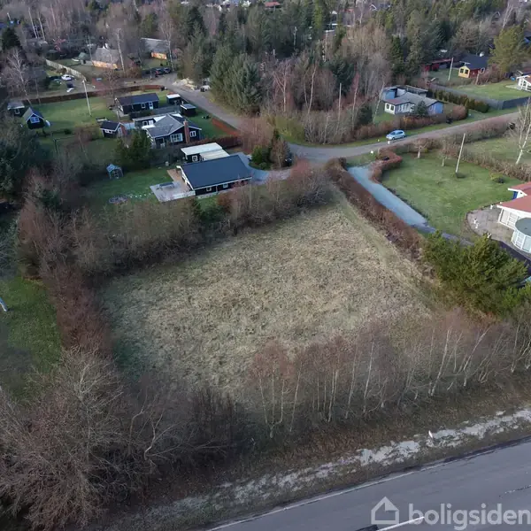 Græsbevokset tomt ligger omkranset af træer og buske, med enkelte huse omkring i et villakvarter. En vej løber parallelt foran tomten.