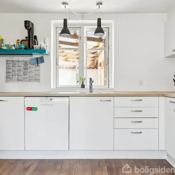 Hvidt køkken med skabsmoduler og indbyggede apparater. To sorte lamper hænger over en træbordplade. Hylde fyldt med dagligvarer. Vindue i baggrunden giver udsyn til gårdhave. Ingen synlig tekst.