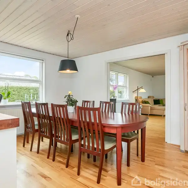 Et spisebord med otte stole står i en lys spisestue. En lampe hænger over bordet. Til højre ses en stue med grøn sofa. En glasvitrine står i hjørnet.