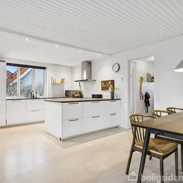 Et moderne køkken med hvidt interiør. Et køkkenbord står midt i rummet med ovn og emhætte. Ved siden af er et spisebord med stole og vase med blomster.